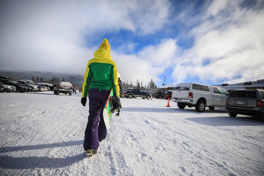 Low-Angle View of a Woman Carrying a Snowboard Through Snowy Parking Lot