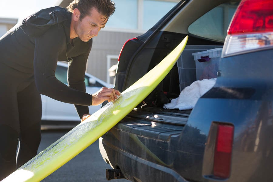 Surfer in a Wetsuit Waxing His Shortboard at the Back of a Car