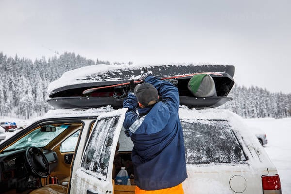 Man Removing a Snowboard from a Roof Carrier at a Parking Lot of a Ski Resort