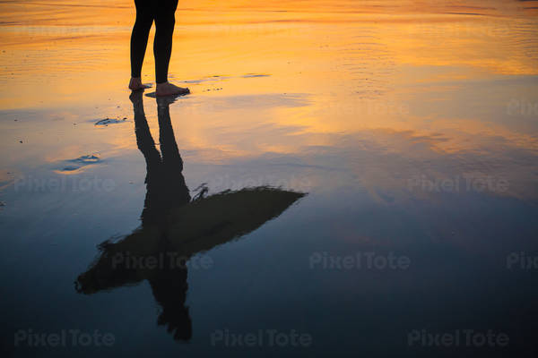 Reflection of a surfer holding a surfboard with a sunset sky behind on a smooth beach sand