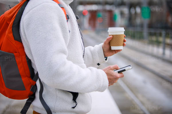 Man Holding a Cup of Coffee Looking at His Phone at a Public Transport Platform