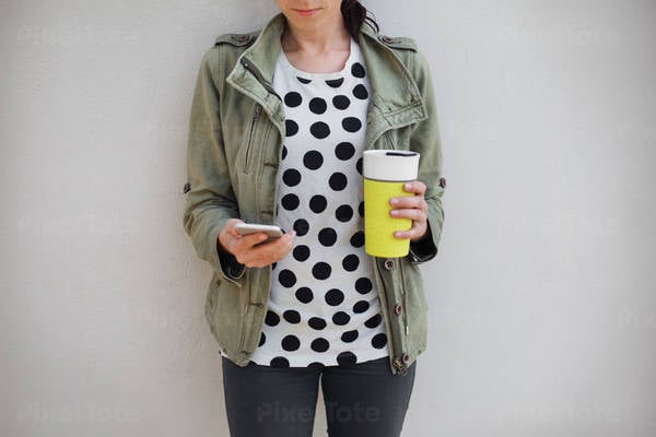 Woman Standing by a Wall Holding a Cell Phone and a Coffee Mug
