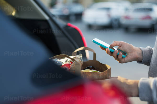 Woman Loading a Bag with Groceries in a Car and Using a Cell Phone