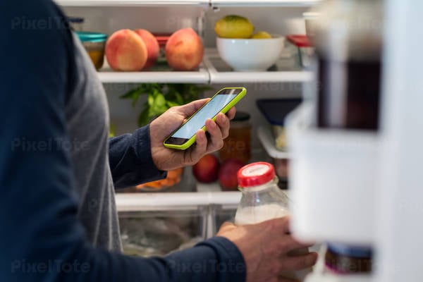 Man Looking at a Grocery Shopping List on a Smartphone at a Refrigerator