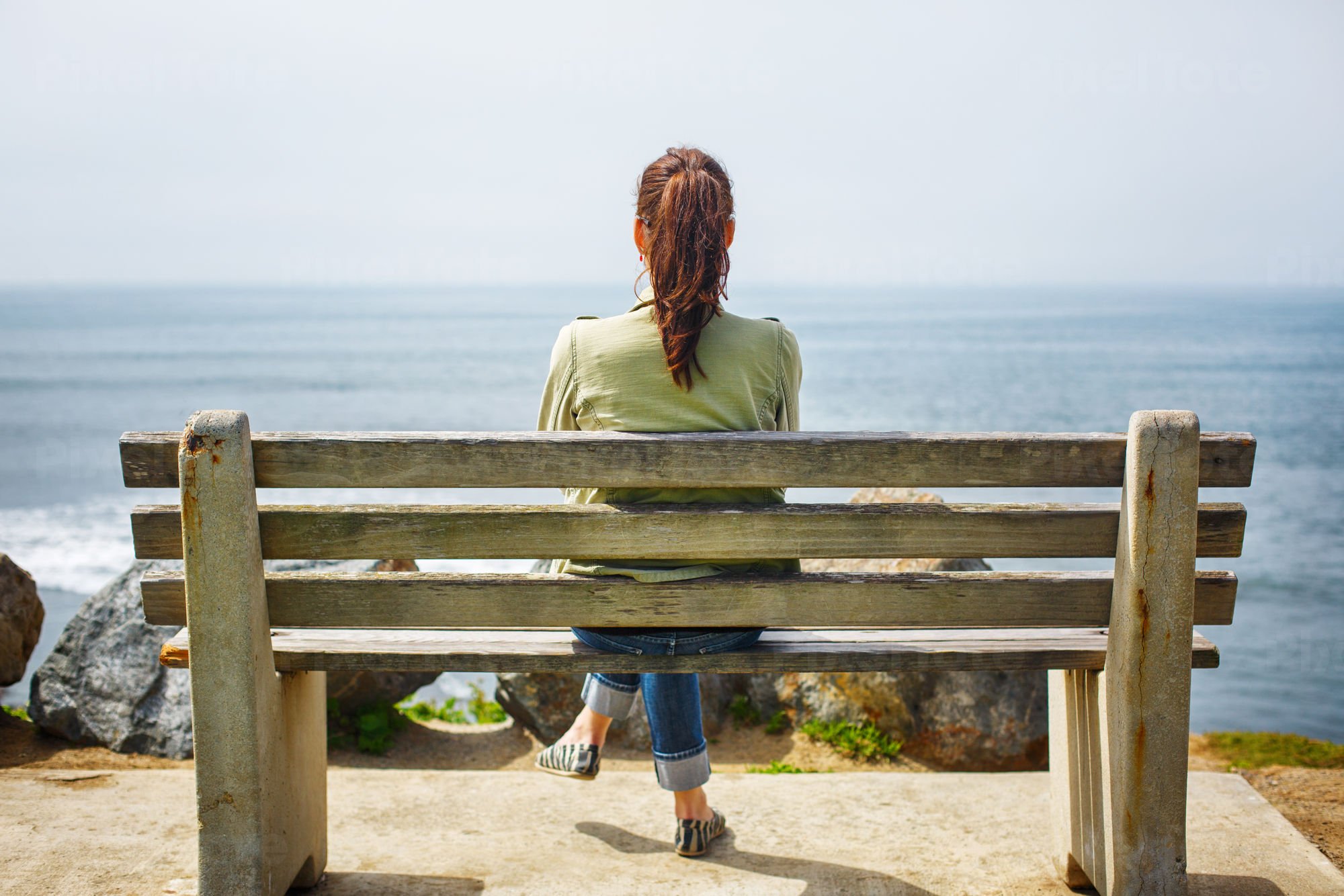 Rear View Of A Woman Sitting On A Bench On A Coast Stock Photo