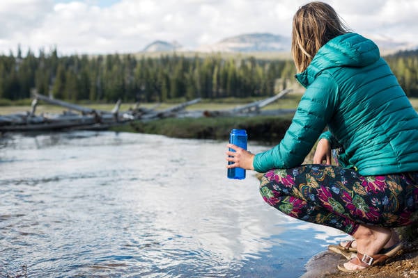 Woman Sitting on a Side of a Mountain Creek and Holding a Water Bottle