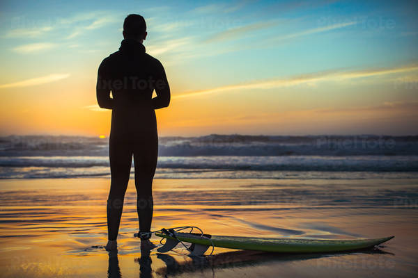 Male Surfer with a Surfboard Standing on a Beach Watching Sunset