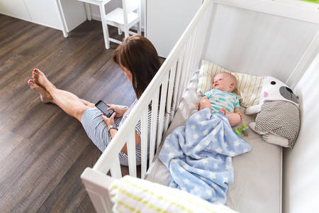 Mom Using a Smartphone Sitting Next to a Crib with Her Baby