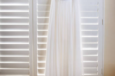 A Wedding Dress Hanging on White Window Blinds at Home