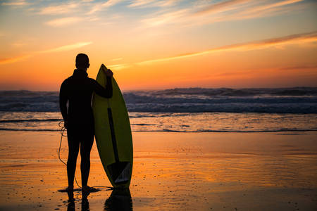 Male Surfer with a Surfboard Standing on a Beach Watching Sunset