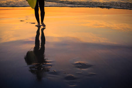 Reflection of a Surfer Holding a Surfboard with a Sunset Sky Behind on a Smooth Beach Sand